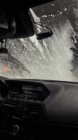 The video shows the interior of a car, specifically the dashboard, during a car wash. The windshield is covered in white foam and water. The car's rearview mirror and a small air freshener are visible hanging from it. On the dashboard is a car stereo system displaying what appears to be audio information in Russian. A phone mount is also attached to the dashboard. The steering wheel is visible on the left side of the frame. The overall lighting is dim, likely due to the enclosed car wash environment.