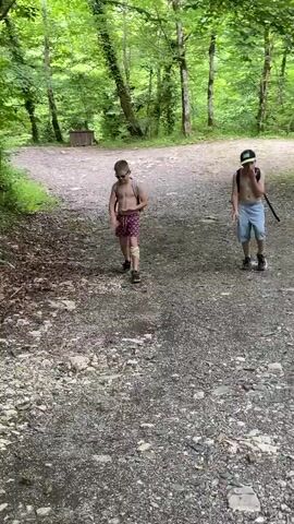 Two shirtless boys are walking on a gravel path in a forest. The boy on the left is wearing patterned shorts and sunglasses, and has a backpack. The boy on the right is wearing light blue shorts and a baseball cap, and is holding a stick. The path is surrounded by trees with green foliage, suggesting it is daytime and likely warm weather. The overall atmosphere appears to be casual and outdoorsy. There is a wooden structure in the background on the left.
