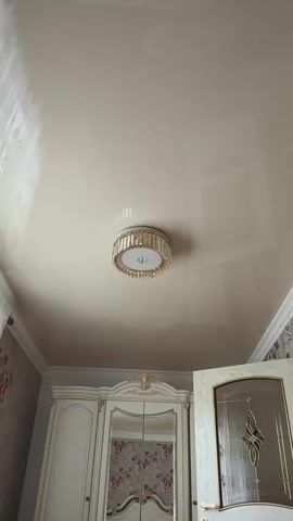 This video showcases a luxurious bedroom interior, focusing on the ceiling and a portion of the room's furnishings. The primary subject is a beautiful, modern ceiling light fixture, circular in design with a white base and a shimmering surround of clear, faceted crystals and gold accents. The light fixture is centrally placed on a smooth, cream-colored stretch ceiling that reflects the light and the room's decor.

Below the ceiling, the top of a grand, ornate wardrobe is visible. It is painted in a cream or off-white color with intricate gold detailing and carving. The wardrobe has large mirrored doors, reflecting a floral wallpaper pattern with pink and burgundy flowers. To the right of the wardrobe, a partially open door with a glass panel featuring a decorative etched design is also visible. The door has a gold handle.

The scene is set indoors, likely in a residential bedroom, given the presence of a wardrobe and the overall aesthetic. The lighting appears to be from the ceiling fixture itself, suggesting it could be daytime or evening, but without external windows clearly visible, the time of day is ambiguous. The reflections on the stretch ceiling suggest a polished and well-maintained environment. There are no people visible in the video. The overall impression is one of elegance and refined taste in the room's decor. The video does not contain any visible text. The location context is Kizilyurt, Russia.