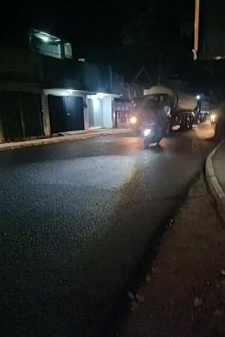 A cement truck is driving down a dark, asphalt road. The truck has its headlights on and is moving towards the viewer. The truck is a light color with a large cylindrical tank on the back. There are some buildings on the left side of the road, and it appears to be nighttime. A motorcycle is also visible in the foreground, illuminated by the truck's headlights. The motorcycle is moving away from the viewer.