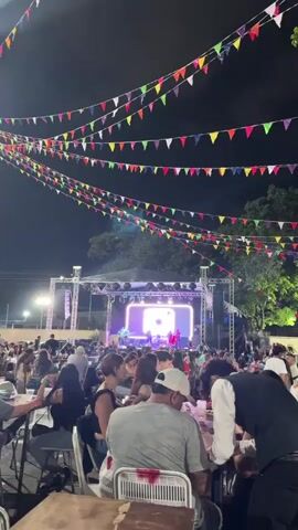 The video shows an outdoor concert at night. People are seated at tables, facing a stage where a band is performing. The stage has a large screen displaying "@labandazohar" and the band's logo. Colorful triangle flags are strung above the audience. The band members are playing instruments and singing. The environment appears festive and lively.
