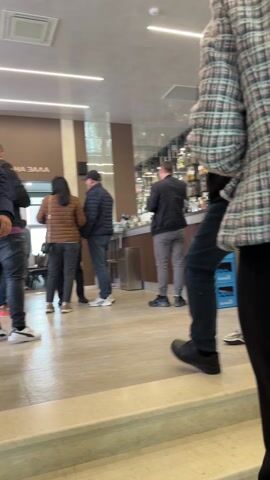 The video shows the interior of a café or restaurant. Several people are walking up a short flight of stairs. One person in the foreground wears a black quilted jacket and jeans. A woman in a black and white patterned jacket and black pants follows. Other people are at the bar, including staff, who appear to be taking orders. The décor includes a painting with music-themed imagery and candelabras. A section of the counter is decorated with artificial greenery. There are shelves stocked with alcohol behind the counter.