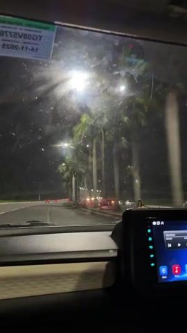 The video begins with a close-up view of a car's dashboard and windshield at night. Raindrops are visible on the windshield, blurring the scene outside. Through the windshield, a line of palm trees can be seen illuminated by streetlights. The road ahead is also lit by streetlights and the headlights of other vehicles, suggesting traffic. Orange traffic cones are placed along the side of the road. The car appears to be moving forward. A digital display screen is visible on the dashboard, showing what looks like music playback controls. The overall atmosphere is dark and wet, indicating a rainy night drive.