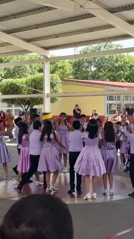 A group of young people are dancing in a circle outdoors under a covered area. The dancers, appearing to be around elementary or middle school age, are dressed in matching light purple dresses for the girls and white shirts with black pants for the boys. Some are holding hands as they move in a circle, while others are in the center of the circle interacting. In the background, more people are gathered, some watching the dancers, and others standing in groups. There are buildings and trees visible in the distance, and what appears to be a school or community center setting. Speakers are set up near the dancing area. The overall atmosphere seems to be festive and celebratory.