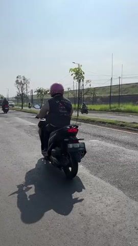 The video shows a person riding a black motorcycle on an asphalt road during the daytime. The rider is wearing a dark vest and a pink helmet. The motorcycle has a license plate visible. The road appears to be a multi-lane road with a grassy median and a fence in the background. There are other motorcycles visible in the distance on the same road. The sky is clear and bright, suggesting sunny weather. The overall scene depicts a typical daytime motorcycle ride on a public road.
