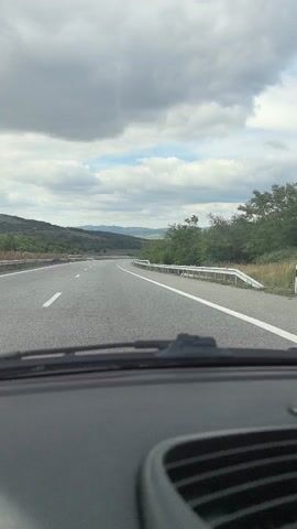 The video shows a first-person perspective of driving on a highway. The road is asphalt with white lane markings. On the right side of the road, there is a metal guardrail. To the right of the guardrail, there are trees and bushes. In the distance, there are rolling hills and mountains. The sky is cloudy with some patches of blue. The dashboard of the car is visible in the foreground.