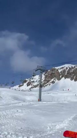 A person is standing on a snowy ski slope on a sunny day with patchy clouds, looking up at a chairlift in operation. Several chairs are occupied as they move up the mountain.  A snow-covered mountain with rocky outcroppings rises behind the lift.  The foreground features a Burton snowboard lying in the snow.  The atmosphere is bright and appears cold.