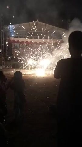 A video shows a pyrotechnic display taking place at night in front of a stage. Bright sparks erupt from the ground, creating a brilliant light and a spray of hot embers. Several people, including a child in a hooded jacket, are visible in the foreground, silhouetted against the light of the fireworks. The stage in the background is illuminated and appears to be set up for a performance. Smoke from the fireworks hangs in the air, obscuring the details of the stage and the sky. The ground is dark and appears to be made of dirt or gravel. The overall atmosphere is one of celebration and excitement.