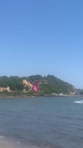 A kiteboarder is shown in the ocean with a large pink kite. The kiteboarder is a silhouette against the bright water and sky. The kite is colorful and appears to be made of fabric. The kiteboarder is in the process of doing a trick and the kite is in the air behind him. The scene is of a beach with trees and a hill in the background. The water is blue and the sky is clear. The overall scene is one of a sunny day with good kiteboarding conditions.