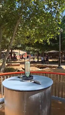 The video shows an outdoor interactive exhibit involving a suspended glass tube. The tube is hanging above a round table with a metal cylinder base. A metal tool rests on the table beside the base. The glass tube is raised and dropped, crashing onto the base. The exhibit is located in a park-like setting with trees, walkways, and other people in the background.  The video pans up to show the rigging supporting the suspended tube, and then back down to the exhibit.