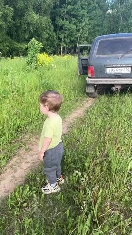 A young boy in a lime green t-shirt and dark grey pants stands on a dirt path in a field of tall green grass. He is facing the left side of the frame, then turns his head to look towards the camera. A dark green SUV is parked on the grass behind him, with its driver's side door slightly ajar. Trees are visible in the background.