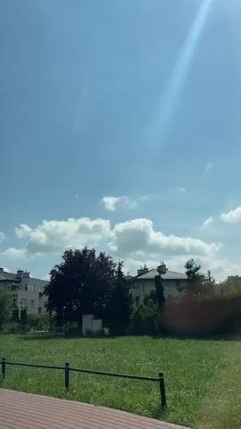 A video shows a view from inside a vehicle looking out at a residential area on a sunny day. The sky is blue with scattered white clouds, and a bright sunbeam is visible in the upper right corner. In the foreground, a wooden deck or surface is partially visible. Beyond that, a green grassy area is present, bordered by a low black fence. Several buildings, appearing to be houses or apartment buildings, are visible in the middle ground, with a large tree with dark red foliage prominently placed in the center-left. The camera is moving horizontally, indicating the video was likely filmed from a moving vehicle. The scene is static with no visible people or actions taking place.