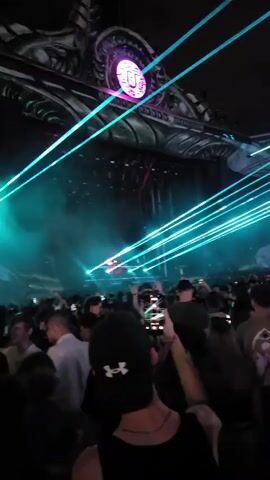 A large crowd of young adults is gathered in a dark, enclosed space. The scene is illuminated by bright green and blue laser beams emanating from a large, ornate structure overhead. This structure, possibly a stage or part of the venue's architecture, features a prominent circular design with a stylized "U" in the center, glowing purple. Many people in the crowd are holding up their cell phones, presumably to record the event. Their faces are mostly obscured by the low lighting and the crowd itself, but a few individuals are visible, looking up at the lasers and the overhead structure. The overall atmosphere appears to be that of a music festival or concert.