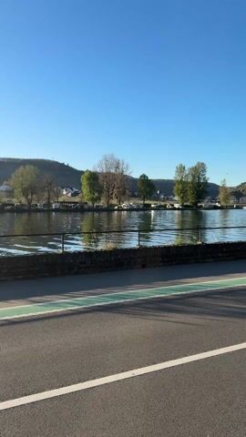 The video shows a road beside a river on a sunny day. The road has a bike lane marked with green paint, and a white line separates the bike lane from the road itself. A row of parked cars is on the other side of the bike lane. The river is bordered by a low brick wall, and beyond that is a mountain covered in greenery, with some houses and vineyards visible. The sky is clear and blue.