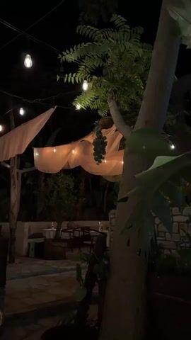 The video shows an outdoor seating area at night. String lights are hung overhead, illuminating sheer fabric draped over a pergola. Tables and chairs are set up on a stone patio, surrounded by greenery and trees. The scene is dimly lit, creating a relaxed and atmospheric ambiance.