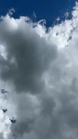 The video shows a close-up, low-angle shot of a blue sky filled with cumulus clouds. The clouds are predominantly white and gray, with some areas appearing darker, suggesting depth and density. The camera is static, looking upwards, and the clouds drift slowly across the frame. The lighting suggests it is daytime with sunlight illuminating the clouds from above. No people, objects, or specific actions are visible. The environment is solely the sky and its cloud formations.