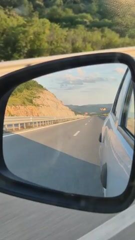 The video shows a view from the side mirror of a car. The mirror reflects a highway with a car driving in the left lane. The highway curves to the right and is bordered by a metal guardrail on the left and a hillside with trees on the right. The sky is blue with some clouds, and the sun appears to be low in the sky, casting long shadows. The video is shot from the perspective of someone in the passenger seat of the car.