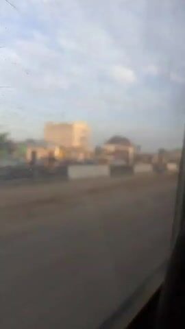 The video shows a view from inside a vehicle looking out at a street scene. The foreground is dominated by the interior of the vehicle, including part of a steering wheel and a side window, which is dusty and smudged. The window frames the view of the street. In the middle ground, there is a road under construction or repair, with a dirt and gravel surface. Concrete barriers are placed along what appears to be a median or a separation between lanes. Several cars are parked or stopped behind these barriers. On the other side of the barriers, there are buildings, some of which appear to be commercial establishments with signs. There are also utility poles with streetlights. A single person, a male wearing a light-colored shirt and dark pants, is standing in the middle of the street, slightly to the right of the frame, in a relaxed posture. In the background, there are more buildings and the sky is visible with scattered clouds. The lighting suggests it is either early morning or late afternoon. The overall impression is a street scene in a developing area, possibly undergoing some infrastructure work.