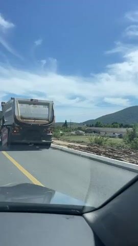 The video captures a view from inside a vehicle driving on a road under construction. A large white dump truck is visible in front. Traffic controllers in high-visibility vests and hats are present. The environment appears to be rural, with hills and vegetation in the background. The sky is blue with scattered clouds. The road has a yellow line marking the lane. Signs and construction barriers are also present. The dump truck has the number 693 visible on its side. The video shows movement as the vehicle drives forward, passing the construction workers and the dump truck.