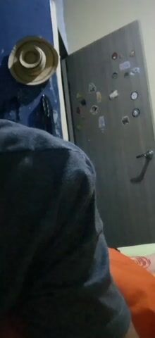 Here is a concise description of the video:

The video shows the interior of a room with a bed, a door, and a hat hanging on a wall. The camera is focused on a person wearing a dark-colored top, with the background consisting of a gray door adorned with various stickers and a dark blue wall with a hat hanging from it. The bed has an orange-colored bedspread.