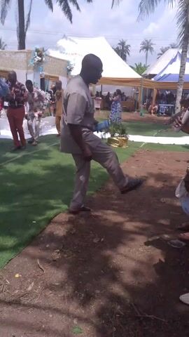 Here's a description of the video:

The video shows a man in a gray suit stepping into a bucket held by another man with a microphone in his hand, apparently at an outdoor event. The man with the microphone, who is wearing a patterned shirt and black pants, seems to be emceeing or commentating on the event. The man in the gray suit has his left foot in the bucket, but it appears to be almost empty. 

In the background, there are tents set up and people milling around. The event appears to be taking place on grass and dirt with palm trees in the background.  A bit of music can be heard.