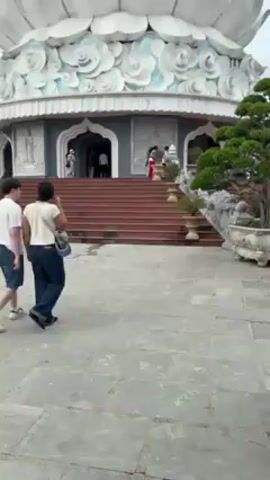 The video shows a large, white, statue of a woman standing on a lotus pedestal, with a building at its base. Two people walk in the foreground on a paved area. The statue is the primary focus, taking up most of the frame. The sky is overcast. The building at the base of the statue has a staircase leading to its entrance. There are small plants and decorative pots around the base of the statue and building. Several people can be seen entering the building.