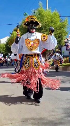 A person in a black mask and elaborate costume, adorned with gold and pink fringe, dances in the street. They wear a white shirt with puffy sleeves, a decorated belt, and black pants and shoes. The person holds a decorated object in their right hand and a gold accessory in their left. In the background, other costumed individuals, onlookers, trees, and buildings are visible under a bright, sunny sky. The scene appears to be a street parade or festival.
