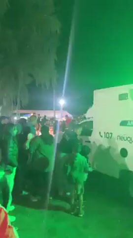 At night, a white ambulance with green and black markings stands near a crowd of people at an outdoor event. The vehicle has "107" and what appears to be Thai script on it, as well as the word "Ambulance". The scene is lit by street lamps and flashing green, red, and blue lights from the ambulance. People are walking around and standing near the ambulance, with one man wearing a security vest. A music stage with blue lighting can be seen in the background. The camera moves around the ambulance.