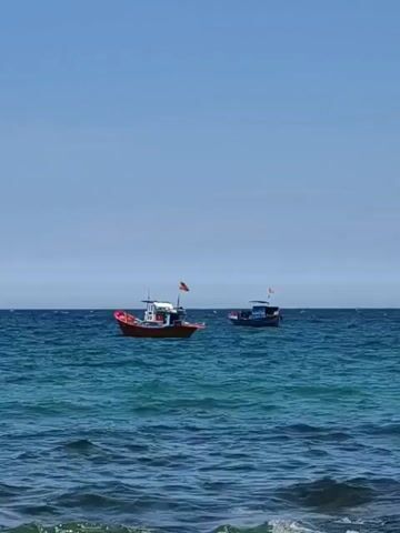 The video shows a wide shot of the ocean under a clear blue sky. There are several small boats on the water, some appearing to be fishing boats. The water is a deep blue-green, with gentle waves. In the distance, a hazy outline of land or islands can be seen on the horizon. The video appears to be filmed from the shore or a slightly elevated position.
