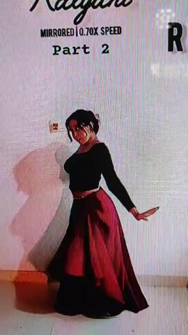 A person in traditional Indian attire, a dark top and a flowing red and black skirt, is performing a dance. The dance appears to be a classical Indian dance form, with graceful movements and expressive hand gestures. The person's hair is tied back, and they are wearing minimal jewelry. The background is a plain white wall with a shadow cast on it, suggesting stage lighting. Text overlays on the video indicate it's "Kalyani MIRRORED | 0.70X SPEED Part 2," suggesting it's a mirrored version of a dance performance slowed down for instructional purposes.