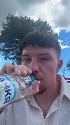 A man is drinking from a water bottle. He is wearing a light-colored shirt. Behind him, there are trees and a blue sky with clouds. There are also some large, colorful letters visible in the background. The man has dark hair with a bowl cut and a goatee.