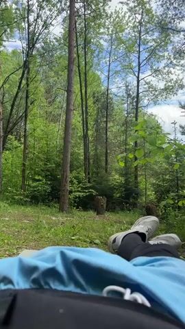 The video shows a first-person perspective of someone lying on their back in a forest, possibly on a sleeping pad or similar surface, with their legs extended towards the camera. The person is wearing dark pants and light-colored shoes. A blue surface, possibly a sleeping pad or tarp, is beneath them. 

The environment consists of a lush green forest with tall trees, green foliage, and grass. Sunlight filters through the trees, creating dappled light. A tree stump is visible in the middle distance. The sky is partially visible through the trees and appears bright with some clouds. The overall impression is one of relaxation in a natural setting.