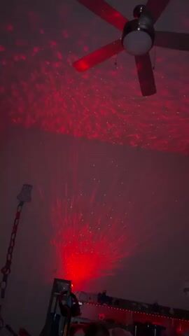The video shows a ceiling fan in a room. The room is illuminated by a light projector that casts red and green light patterns on the ceiling and walls, resembling water waves and a burst of light. There is also a lamp visible on the left side of the screen, and what appears to be a bed or furniture in the background. The light projector itself is visible at the bottom of the frame, emitting the red light. The scene appears to be set up for a calming or atmospheric mood.