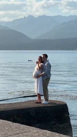 The video shows a pregnant woman in a white dress standing on a concrete pier with her partner, who is in a striped shirt and khaki pants. They are embracing by the water with mountains in the background. The woman is holding her belly, and the man has his arm around her. There are two people on a paddleboard in the water behind them. The sky is partly cloudy.