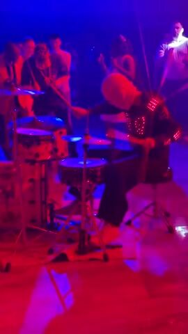A person wearing a white hood and a black jacket adorned with red lights is playing a drum set. Red laser beams are visible throughout the scene. Several other individuals are in the background, some appearing to be watching or dancing. The environment looks like an indoor venue, possibly a club or event space, with some stage lighting and smoke.