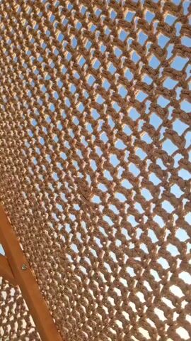 The video opens with a close-up view of a woven net, possibly from a hammock or a shade structure. The net is made of thick, light-colored rope and forms a diamond pattern. Through the holes in the net, a bright sun is visible in a clear blue sky, casting a shimmering reflection on the water below. As the camera moves, the perspective shifts to reveal more of the environment. The sun appears to be setting or rising over a calm body of water, with a few dark rocks or small islands visible in the distance. The surface of the water is slightly rippled, and the sunlight creates a bright, bokeh effect on its surface. A large, white object, possibly an umbrella or the canopy of a boat, partially obscures the upper right portion of the frame. The overall impression is of a peaceful, sunny day by the sea.