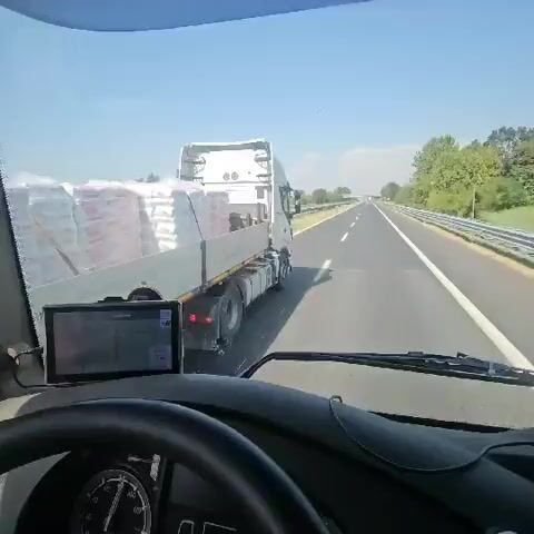 The video shows a first-person view from inside a truck cabin. The driver's steering wheel is visible in the foreground, along with part of the dashboard. A GPS device is mounted on the dashboard. The truck is on a highway, with other vehicles visible ahead. The truck is carrying a load of large white bags on its trailer, which are covered with plastic. The weather appears to be sunny and clear. The truck is in motion, traveling down the highway.