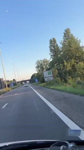 The video shows a view from the driver's seat of a vehicle traveling on a highway. A highway sign indicating "Willebroek 1000m" is visible on the right side of the road. There are other vehicles on the highway, and overhead bridges can be seen in the distance. The sky is clear and blue. The sun appears to be low, casting long shadows on the road. There are trees and other vegetation on the sides of the highway. The video captures the perspective of someone driving, with the dashboard and windshield visible at the bottom and top of the frame, respectively.