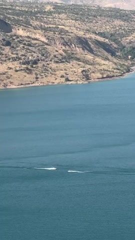 The video captures an aerial view of a large body of water, likely a lake or reservoir, with a mountainous and arid landscape surrounding its shore. The water is a deep blue-green color, with gentle ripples indicating a calm day. Two small personal watercraft, possibly jet skis, are visible on the water, leaving white wakes as they move. The shoreline features rugged, tan-colored hills sparsely covered with dry vegetation and some scattered trees. In the distance, more prominent mountains rise, their peaks hazy and bathed in sunlight. The overall impression is a vast, natural landscape with recreational activity on the water.
