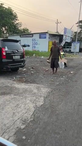 The video shows an outdoor scene with an unpaved road covered in debris. A black SUV is parked on the left side of the frame. In the center of the frame, a man wearing a dark shirt and shorts is walking towards the camera, carrying a brown object in his left hand and a plastic bag in his right. Behind him, a woman wearing a green shirt and dark pants is riding a motorcycle, with another person behind her. A yellow building with a sign reading "SILVIA PHONE" and advertisements for mobile data plans is visible in the background. The sky above is a pale orange, suggesting it is either dawn or dusk.