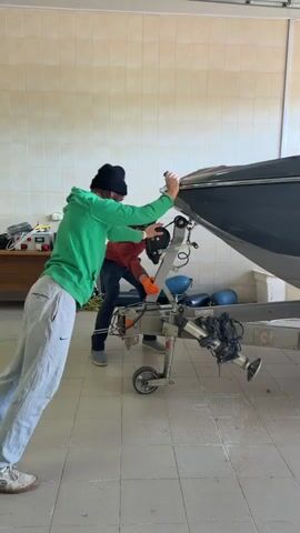 Two men are seen moving a gray-blue boat on a silver trailer out of an indoor garage or storage area. The garage features light-colored tiled walls and a matching tiled floor. Bright sunlight streams in from the open garage door at the far end, indicating it is daytime.

One man, wearing a green hoodie and gray sweatpants, is positioned at the front of the boat, pushing it forward. The second man, dressed in an orange long-sleeved shirt and dark pants, is operating the front jack stand and wheel mechanism of the boat trailer, guiding its movement. The boat, with its hull reflecting light, slowly glides forward on the trailer.

The registration number "TB4406RUS69" is visible on the side of the boat's hull. The brand name "CENTURION" can also be seen on the trailer's frame. An audible mechanical hum accompanies the movement of the boat. The activity appears to be the careful maneuvering of the boat and trailer, likely in preparation for transport or storage. The location is in Shchelkovo, Russia.