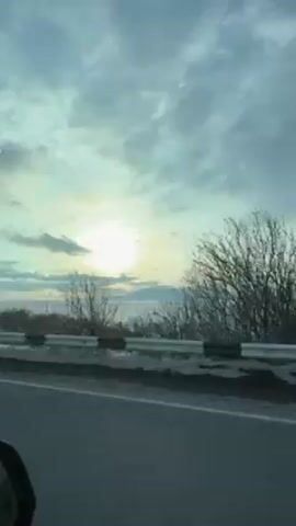The video shows a view from a car driving down a highway. On the right side of the road, there is a guardrail with snow behind it. The road is gray. The sky is cloudy with a sun low on the horizon. There are barren trees on the right side of the road. A white van drives past. There is music playing.