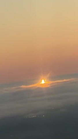 The video shows a sunrise from above clouds. The sun is a bright, glowing orb peeking over a layer of clouds, casting golden light and rays. The sky is a gradient of orange and pink. Below the clouds, faint lights suggest a landscape or city.