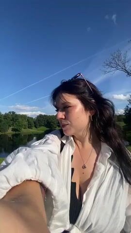 A woman with dark hair and sunglasses perched on her head, wearing a white unbuttoned shirt over a dark top and a patterned skirt, takes a selfie with her right arm extended. The background shows a park with a body of water on the left, green grass, trees, a pathway, and a light-colored building in the distance. The sky is blue with scattered clouds and the sun is visible, creating lens flare. The woman smiles and looks towards the camera in some frames, and looks away in others, appearing to walk along the pathway. Other people can be seen in the distance walking on the path. The video is a series of short clips, suggesting it was filmed during a walk.