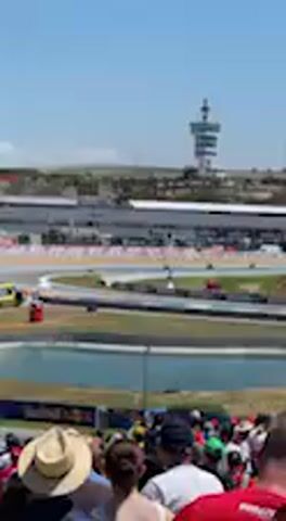 The video captures an outdoor racing event under a bright, clear sky. In the foreground, a partial view of spectators shows heads and shoulders, with individuals wearing hats and casual clothing, indicating a daytime event. The main action unfolds on a race track in the mid-ground, where multiple racing vehicles are visible, appearing as streaks of color due to their speed as they navigate a wide, sweeping turn. The track features asphalt, marked white and red kerbs, and green run-off areas. Adjacent to the track, a blue body of water is visible. In the background, a distinctive white control tower stands tall against the horizon, overseeing the circuit. The general atmosphere is lively, with the audible roar of engines suggesting an ongoing race. This event is taking place at a race circuit in Jerez de la Frontera, Spain. No discernible text is visible on the track, buildings, or banners.
