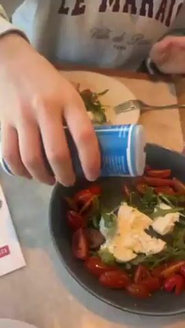 A hand is sprinkling salt from a blue container onto a salad in a dark grey bowl. The salad contains quartered red tomatoes, arugula, and a white creamy cheese. A plate with a piece of bread and a knife and fork sit next to the salad. The table is marbleized. The person sprinkling the salt is wearing a grey sweatshirt with the words "Le Marais Ville de Paris" on it. A basket with a red and white checkered lining is on the table.