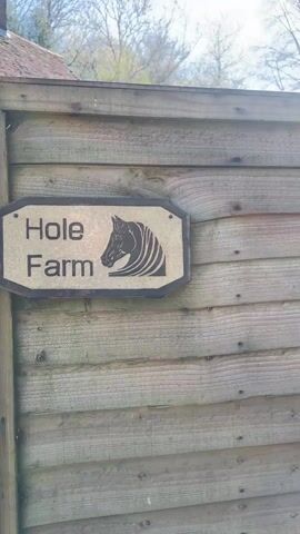 The video shows a rustic sign that reads "Hole Farm" with a horse emblem on a wooden fence, then pans to a man operating a cement mixer near a gate. The camera continues to reveal a large, old-fashioned stone house with dark wooden beams and a brown tiled roof, surrounded by a green lawn and trees. A Land Rover Defender is parked near the house. The scene is bright and sunny.