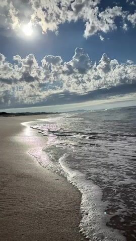 The video shows a sunny beach with gentle waves.  The sky is blue with scattered puffy white clouds. The sun is shining brightly through the clouds, reflecting off the wet sand and the water's surface. Small waves are rolling onto the shore, with foam visible.  The sand appears to be light brown. A strip of greenery is visible in the distance along the shoreline.
