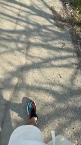 A first-person view video shows someone walking on a cracked asphalt road. The person is wearing white shorts and colorful running shoes. The road is lined with trees casting shadows, and there are buildings visible in the distance. The sky is blue. The video appears to be filmed during the day.