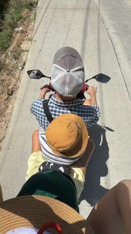 The video shows a first-person perspective of someone riding on the back of a motorcycle. In front, another person is driving, wearing a blue and white checkered shirt and a grey baseball cap. A child is seated between the driver and the passenger, wearing a striped shirt and a tan bucket hat. The passenger is wearing a straw hat. The motorcycle is on a paved road with a grassy and rocky area to the left. The lighting suggests it is daytime with sunlight casting shadows.